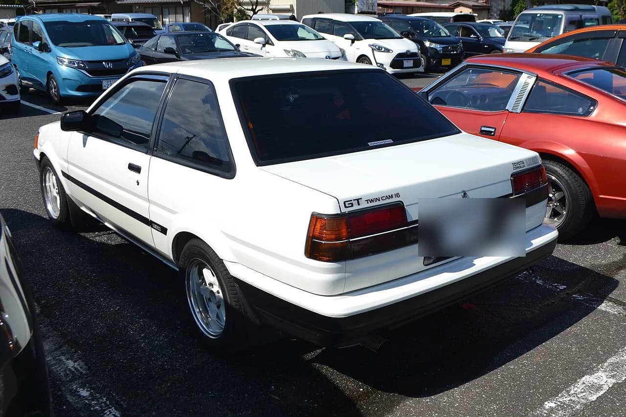 A white Toyota Corolla Levin AE86 GT parked at a car meet, rear three-quarter view (license plate blurred)